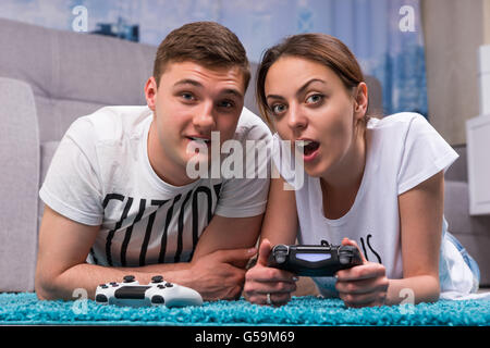 Emotional young couple lying on a rug in their living room playing video games looking towards the camera and pointing with fascinated expressions. Stock Photo