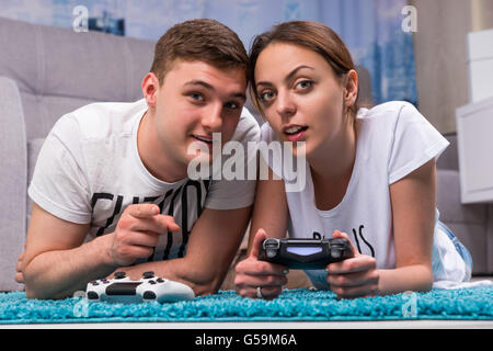Young couple lying on a rug in their living room playing video games looking towards the camera and pointing with fascinated expressions. Stock Photo
