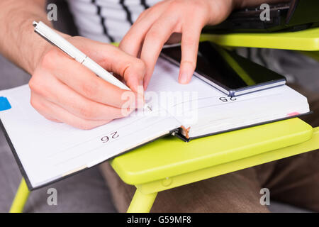 Close up of man's hand writing  making appointments in diary lying on the laptop table doing his business from home sitting on a sofa holding a pen. Stock Photo