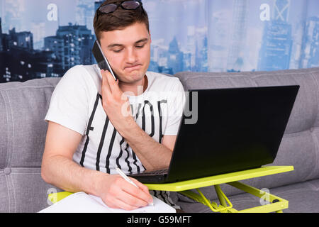Harassed man working from home sitting on a sofa talking on a mobile phone while writing notes and using a laptop computer Stock Photo