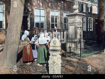 Auf der Insel Walcheren liegt noch das ebenfalls recht stille, aber trauliche Domburg. Die Häuser sind aus Backstein mit bunten nach außen aufgehenden Fenstern. Auch die Straßen und Wege sind wie vielfach mit hochkantstehenden Backsteinen gepflastert. Am Brunnen hat sich eine kleins Gesellschaft versammelt, die in den Holzschuhen lustig dahergeklappert ist. Der Brunnen ist an drei Seiten von einem Gitter umgeben. Im Vordergrund steht ein Wappenstein aus alter Zeit. Volkstrachten, Windmühlen und malerische Architektur geben der holländischen Landschaft ein abwechslungsreiches Gepräge. Netherlan Stock Photo