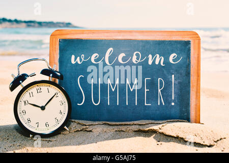 closeup of retro alarm clock and a chalkboard with the text welcome summer written in it, placed on the sand of a beach Stock Photo