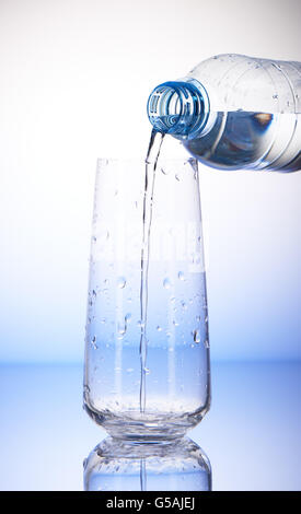 Water pouring from plastic bottle into empty drinking glass with reflection in drops of water on white and blue background Stock Photo
