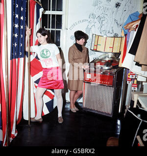 In Pauline Fordham's boutique in Ganton Street, near Carnaby Street. Customer Jackie Binder in a 'with it' blouse and skirt stands beside a dressing room decorated with the Union Jack and the Stars and Stripes. In the background, Jacqueline Ball plays the juke box. Stock Photo