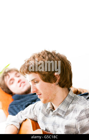 Handsome young man playing guitar for a friend who is resting on a sofa alongside him, close up head and shoulders profile Stock Photo