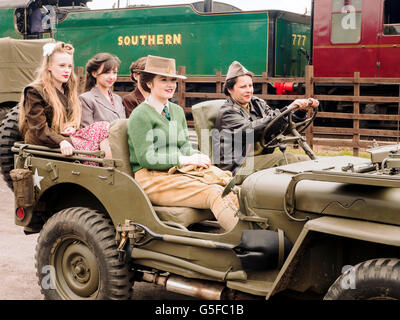 Young women in WW2 (1940's) clothes riding on a jeep with a steam train in the background. NB This is a re-enactment. Stock Photo