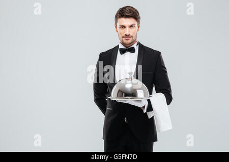 Attractive young waiter in tuxedo holding serving tray with metal cloche and napkin Stock Photo