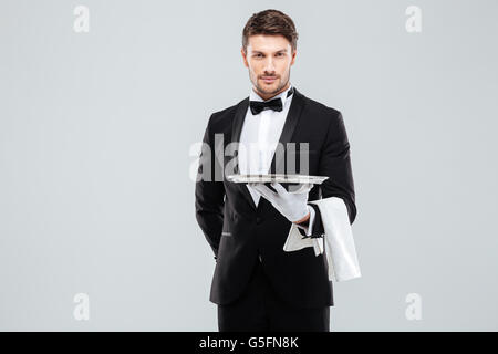 Handsome yong waiter in tuxedo and gloves holding empty tray and napkin Stock Photo