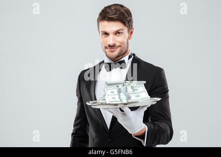 Smiling young waiter in tuxedo and gloves holding tray with money Stock Photo