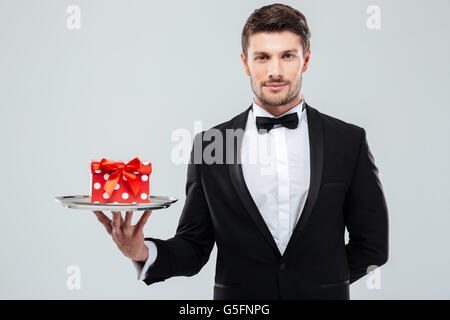 Attractive young waiter in tuxedo with bowtie holding present box on tray Stock Photo