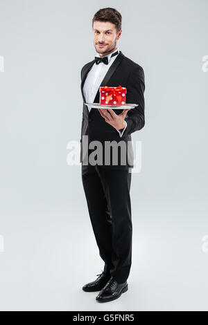 Full length of young butler in tuxedo standing and holding tray with gift box Stock Photo