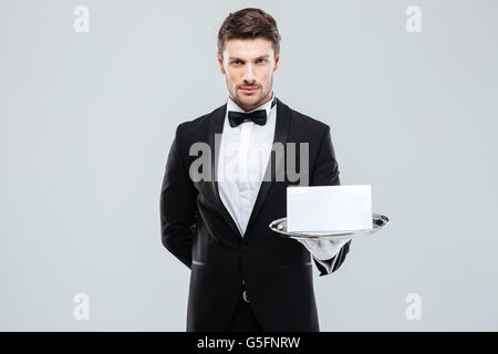 Confident young butler in tuxedo holding blank card on tray Stock Photo