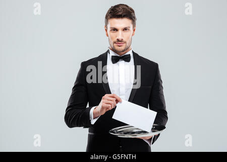 Handsome young waiter in tuxedo holding tray with blank card Stock Photo