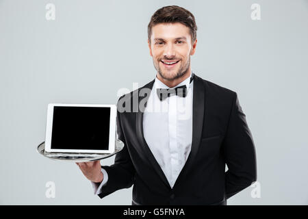 Cheerful young waiter in tuxedo standing and holding blank screen tablet on tray Stock Photo