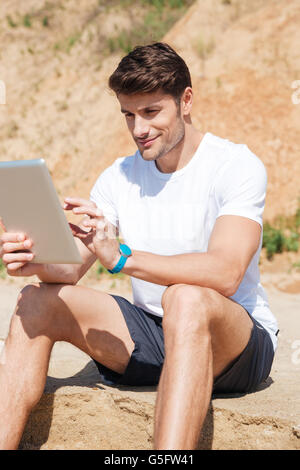 Smiling young man sitting and using tablet on the beach Stock Photo