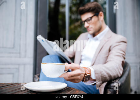 Thoughtful young man in glasses with cup of coffee sitting and reading magazine in outdoor cafe Stock Photo