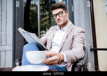 Confident relaxed young man in glasses holding cup of coffee and reading magazine in outdoor cafe Stock Photo