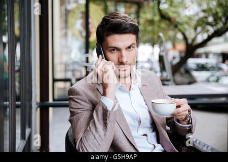 Serious young businessman talking on cell phone and drinking coffee in outdoor cafe Stock Photo