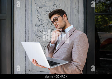 Thoughtful young man in funny round glasses thinking and using laptop Stock Photo