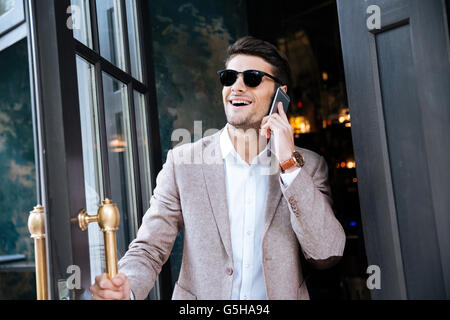 Cheerful young businessman in sunglasses talking on cell phone and walking out from cafe Stock Photo