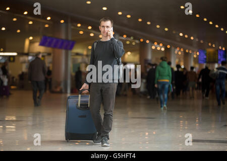 Young handsome man in 20s walking in modern airport terminal, talking on smart phone, travelling with luggage bag Stock Photo