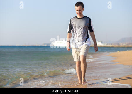 Young Caucasian sporty man walking on the coastline, happy smiling, making splashes, having fun, white yacht on the background Stock Photo