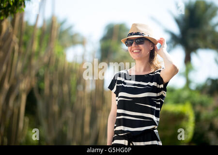 Young woman in straw hat, sunglasses and summer dress walking on tropical resort street, happy smiling Stock Photo