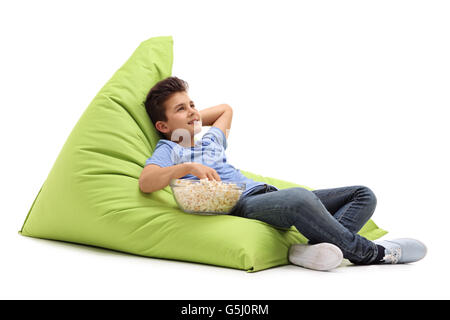 Pensive little boy eating popcorn seated on a green beanbag isolated on white background Stock Photo