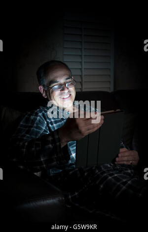 Middle aged man wearing pajamas reading tablet at night in bed looking happy and smiling Stock Photo