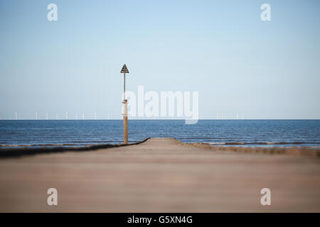 A wooden jetty stretching out into the sea with a wind farm in the distance on the horizon Stock Photo