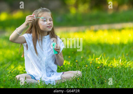Little girl sitting in the grass and make blowing bubbles. Stock Photo