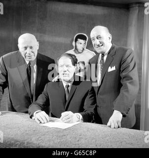 With a life-sized photo model of the man he is to box, Henry Cooper is at the Odeon in Leicester Square, London, to sign the contract for his fight with Heavyweight Champion Muhammad Ali, with the bout to be staged at Arsenal's Highbury stadium on May 21. Watching him are his manager Jim Wicks (left) and promoter Harry Levine (right). Stock Photo