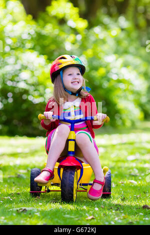 Cute girl wearing safety helmet riding her tricycle in sunny summer park. Kids ride bicycle. First bike for little child Stock Photo