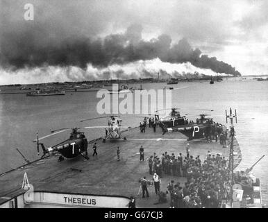 Troops and helicopters on deck of one of the first Royal Navy ships to enter Port Said, Egypt, which is seen shrouded in smoke and fire after its capture. Stock Photo