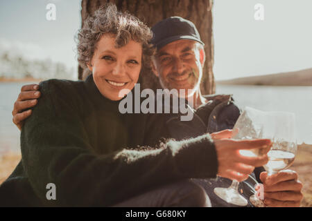 Portrait of loving mature couple sitting together with a glass of wine. Man and woman camping near a lake on a summer day. Stock Photo