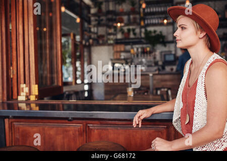 Side view of beautiful young caucasian woman sitting at bar counter and looking away. She is wearing hat waiting for someone. Stock Photo