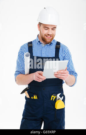 Happy handsome young builder in helmet standing and using tablet Stock Photo