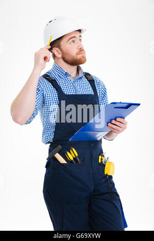 Pensive young builder in helmet thinking and writing on clipboard Stock Photo