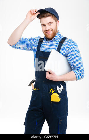 Cheerful attractive young builder in overall and cap standing and holding laptop Stock Photo