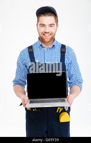 Happy young builder in overall and cap holding blank screen laptop Stock Photo