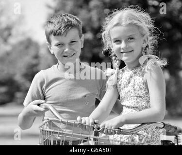 1930s 1940s PORTRAIT SMILING BOY PUSHING BIKE WITH BLOND GIRL PIGTAILS SITTING BY HANDLEBARS BOTH LOOKING AT CAMERA Stock Photo