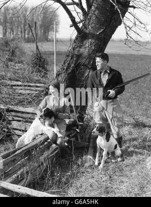 1930s COUPLE IN HUNTING GEAR WITH TWO SPRINGER SPANIEL DOGS WOMAN SITTING ON SPLIT RAIL FENCE MAN STANDING HOLDING SHOTGUN Stock Photo