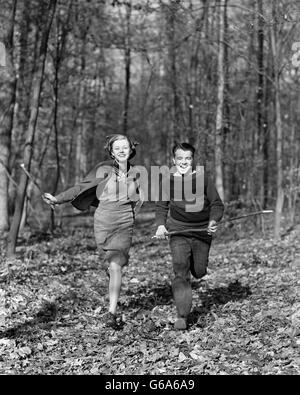 1940s SMILING TEENAGE COUPLE BOY GIRL RUNNING IN AUTUMN WOODS LANDSCAPE Stock Photo
