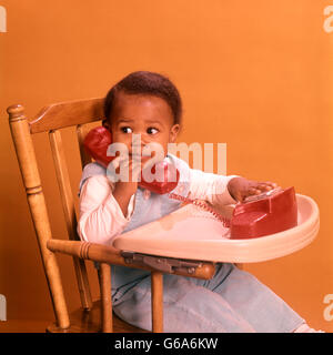 1970s AFRICAN AMERICAN BOY PLAYING WITH TOY TELEPHONE SITTING IN HIGHCHAIR Stock Photo
