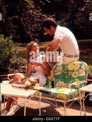 1970s RELAXING COUPLE SITTING ON PATIO FURNITURE WITH DRINKS BOTH WEARING TENNIS TOGS AFTER PLAYING Stock Photo
