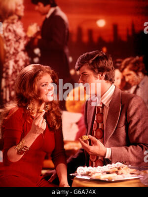 1970s YOUNG COUPLE MAN AND WOMAN EATING FINGER FOOD AT A PARTY Stock Photo