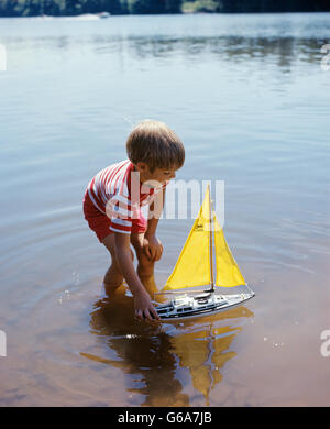 1970s BOY WADING IN POND WATER PLAYING WITH TOY BOAT WITH YELLOW SAILS Stock Photo