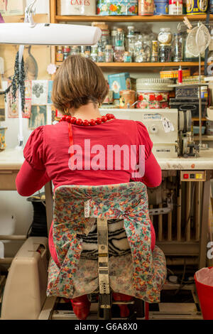 A seamstress in red seated, back turned, and working by a sewing machine Stock Photo