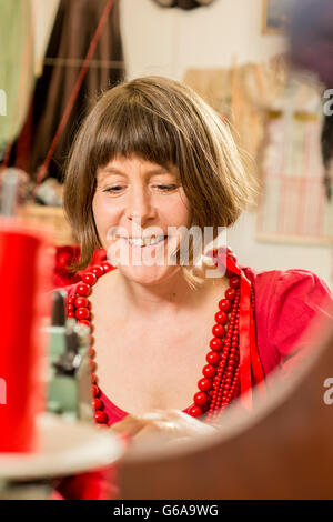 Lady tailor in red dress and necklaces smiling and working on a sewing machine Stock Photo