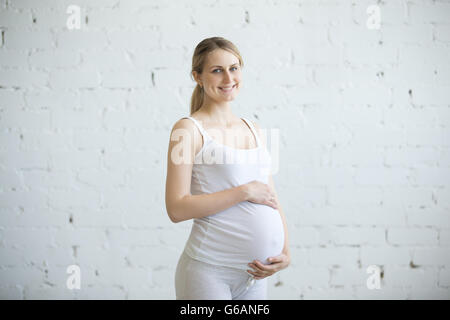 Healthy pregnancy concept. Portrait of beautiful blond young pregnant model looking at camera while caressing her belly Stock Photo